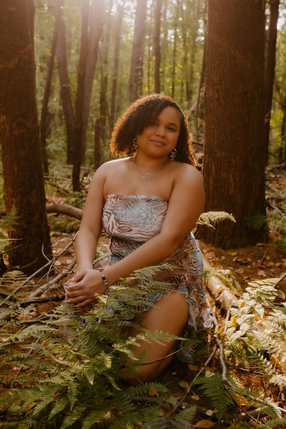 A person in a strapless dress sits among ferns in a sunlit forest during golden hour.