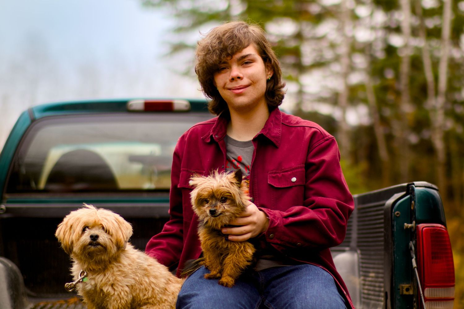 Two small dogs and their companion sit in the back of a pickup truck on a cloudy day.