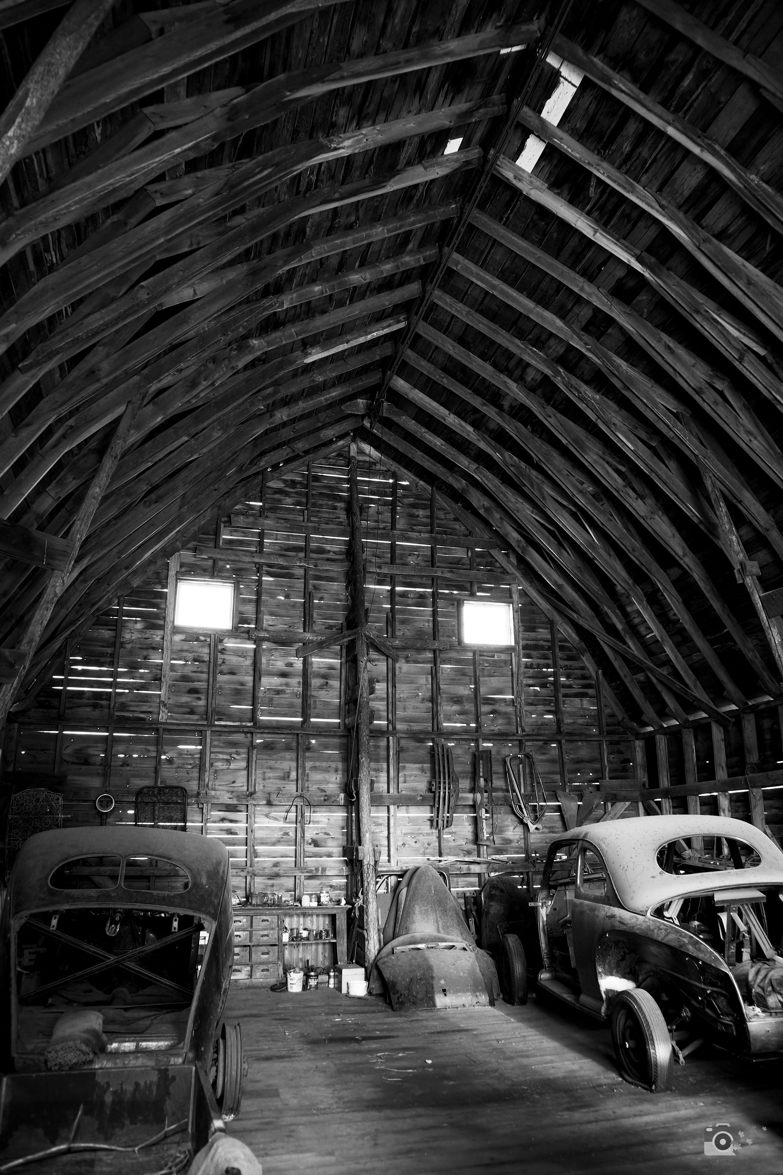 Classic vintage cars parked inside a rustic wooden barn with sunlight streaming through windows in the roof.