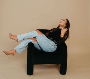 A relaxed pose on a black chair wearing light denim jeans and a dark top against a beige backdrop.