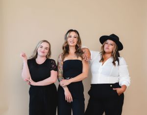 Three women in stylish black and white outfits posing against beige wall.