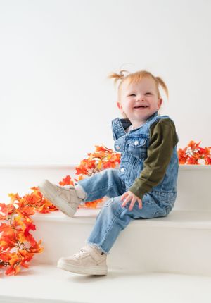 Toddler in denim overalls and green sweater sitting on white steps with orange fall decorations.