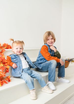 Two children in matching denim outfits sitting on white steps with orange autumn decorations.