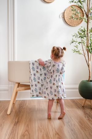 Small child in patterned dress stands facing decorative wall with woven baskets and plant decor.