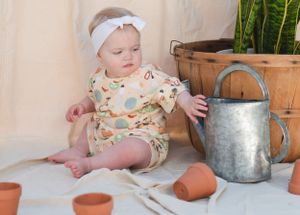 Baby in floral outfit sitting next to vintage watering can and terracotta pots.