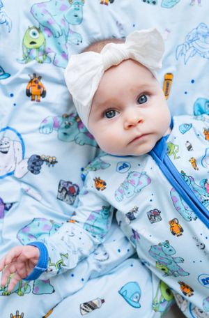 A baby wearing a white bow headband lies on blue patterned bedding with cartoon monster prints.