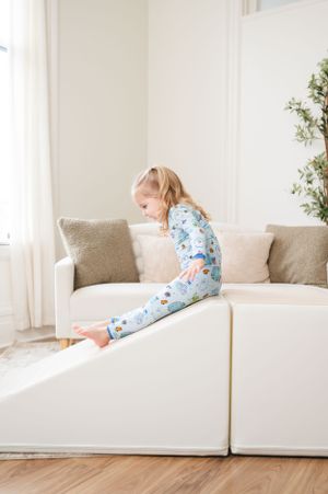 A child in blue floral pajamas plays on a white sectional couch in a bright, airy living room with houseplants.