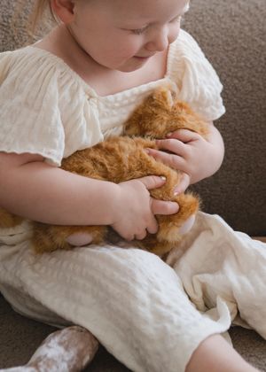 A small child in white clothing gently holds and cuddles an orange tabby cat while sitting on carpet.