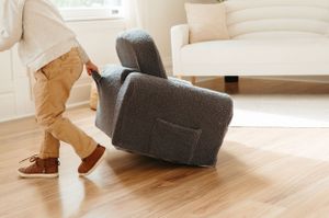 Small legs and feet pushing a grey upholstered chair across hardwood flooring in a bright living room.