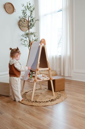 Child stands at wooden easel in minimalist room with woven wall decor and potted plant.