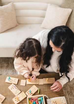 A candid moment on a beige couch shows two people leaning over a wooden table playing with dominoes and colorful markers.