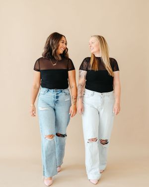 Two women in black tops and ripped jeans smiling at each other against beige background.