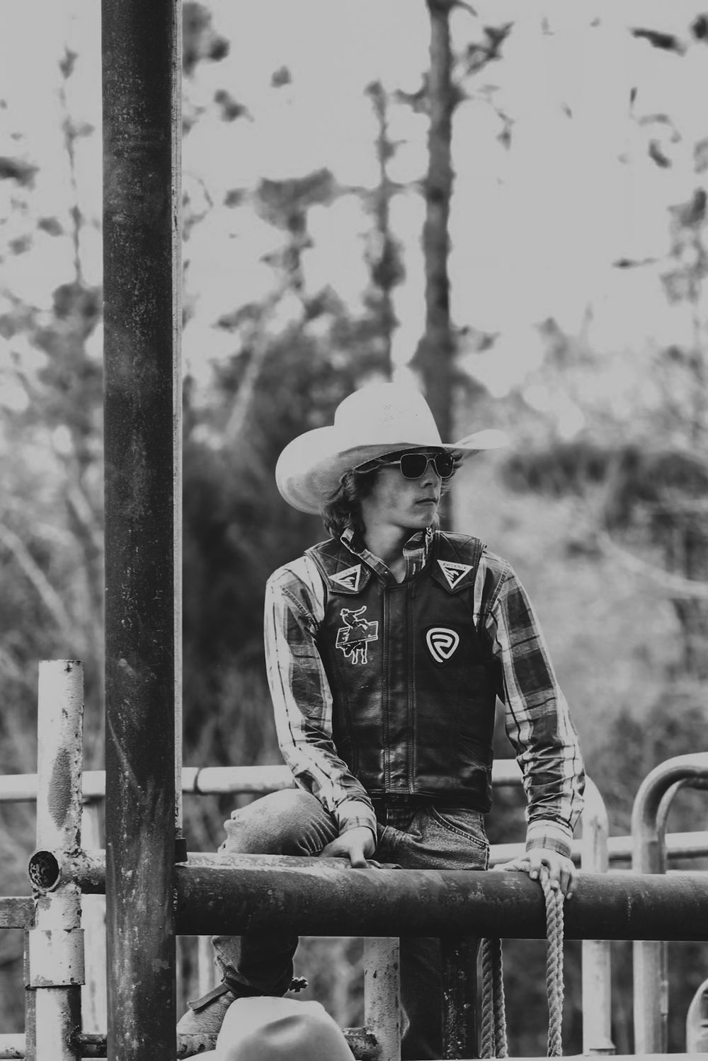 Western-style riders sitting on metal fence at a rodeo arena in black and white.