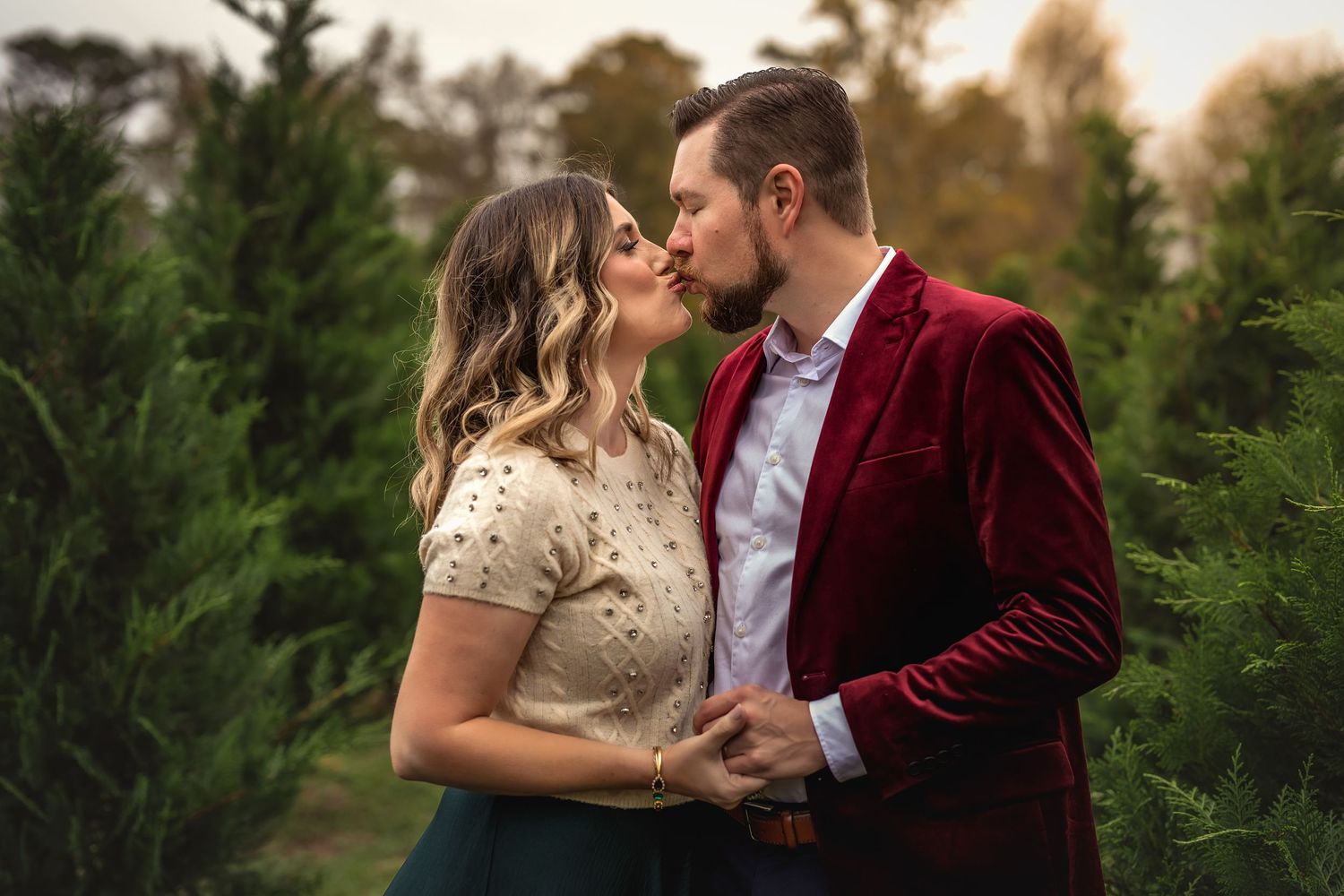 A couple shares an intimate moment surrounded by evergreen trees - she in a bejeweled sweater and he in a red velvet jacket.
