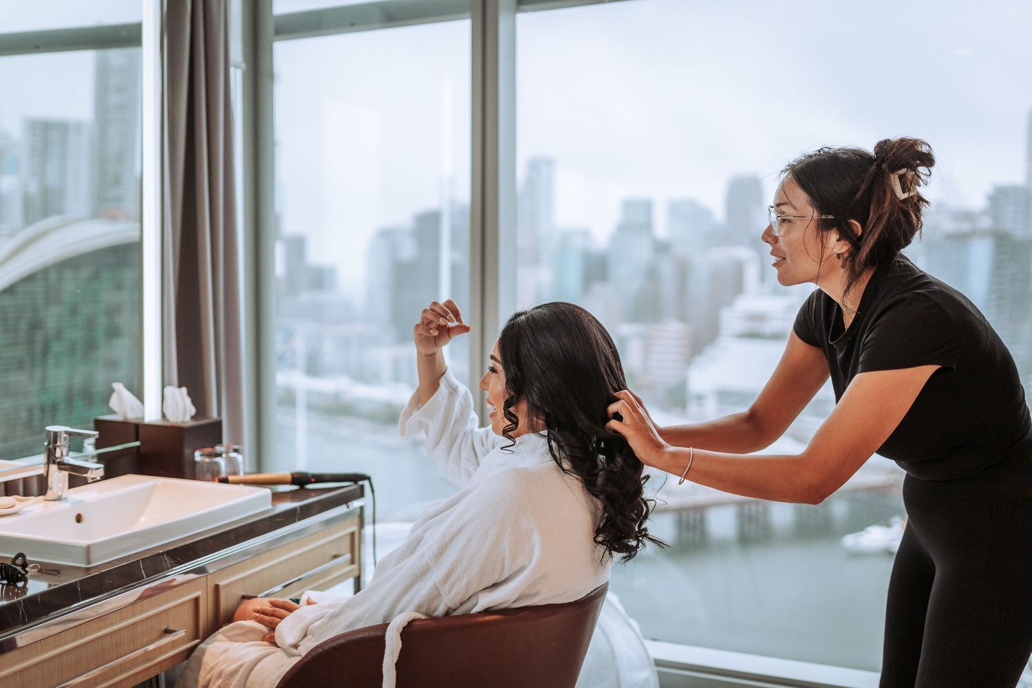 Hair styling session during wedding preparations with city skyline visible through windows.