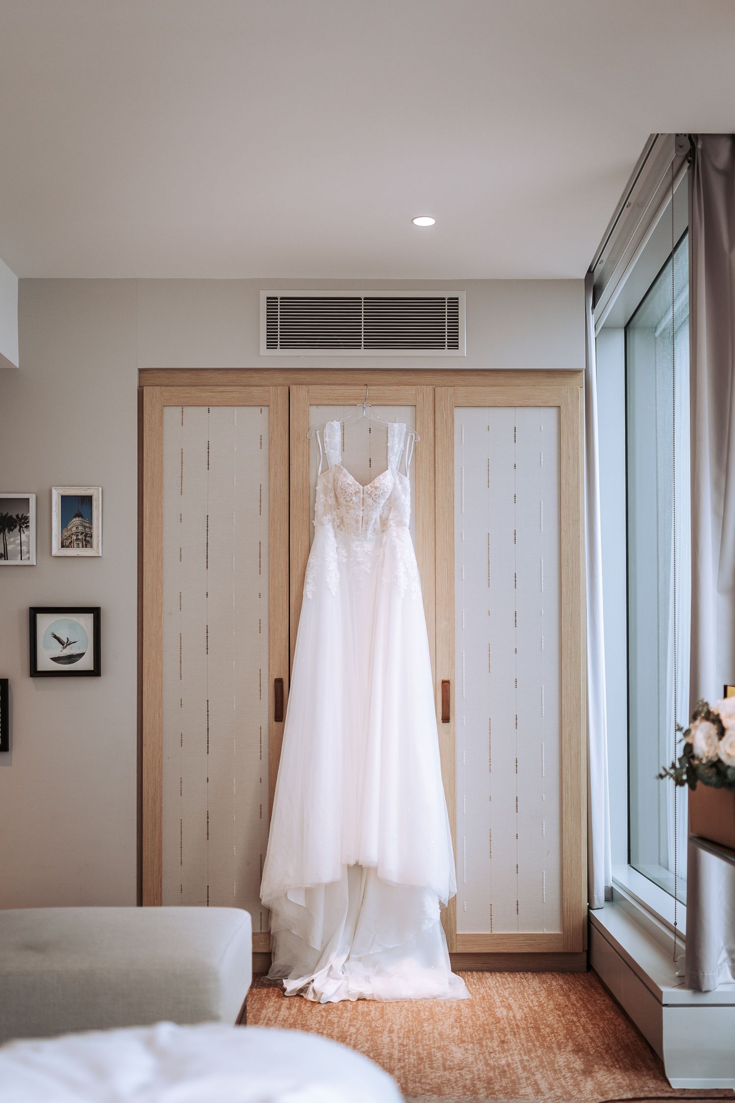 An elegant white lace wedding dress hangs from a closet door in a sunlit room.