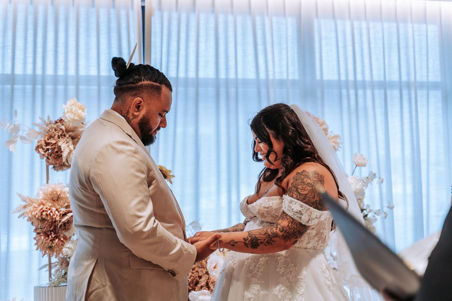 Couple exchanges rings during intimate indoor wedding ceremony with light streaming through sheer curtains in background.