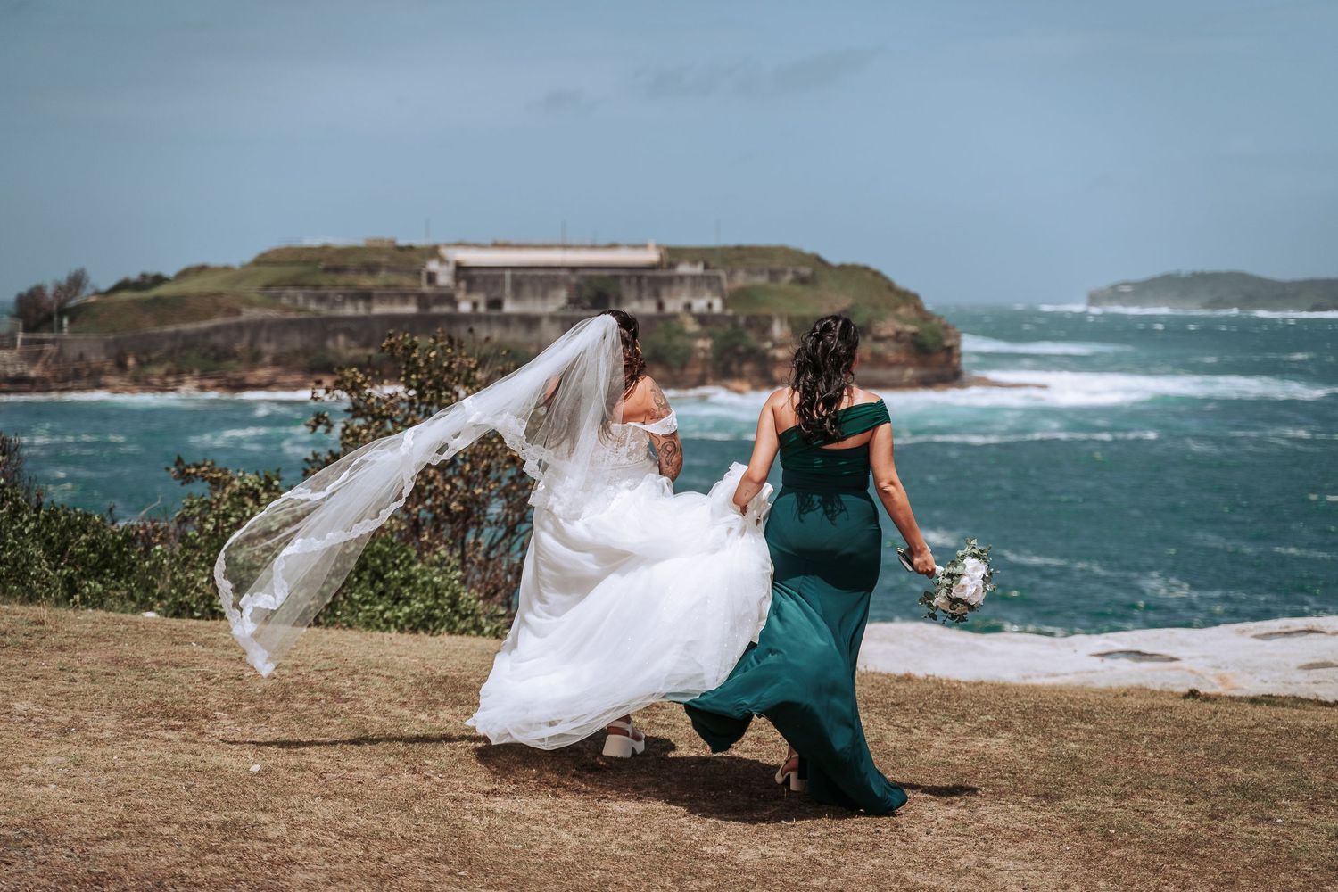 Wedding dress and veil flowing in wind on coastal clifftop with ocean and buildings in background.
