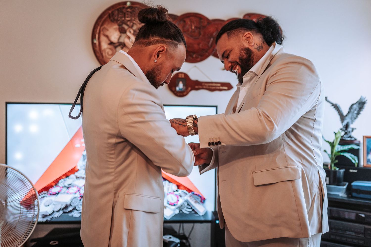 Two people in beige suits helping each other get dressed for a formal occasion.