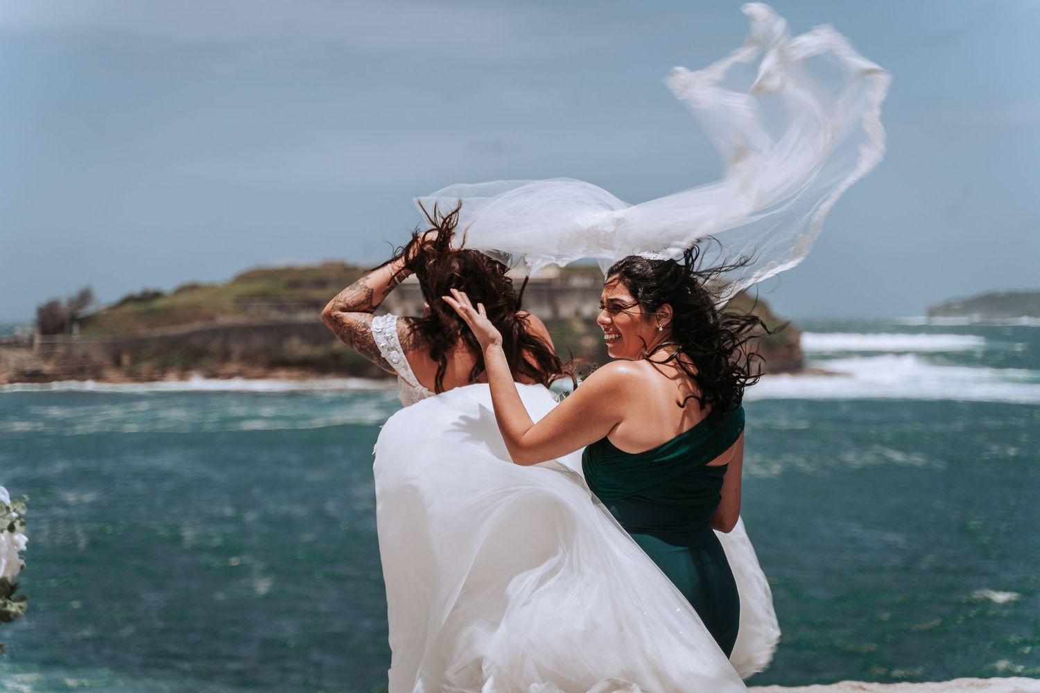 Wedding couple embracing on windy coastal cliff with ocean waves in background.