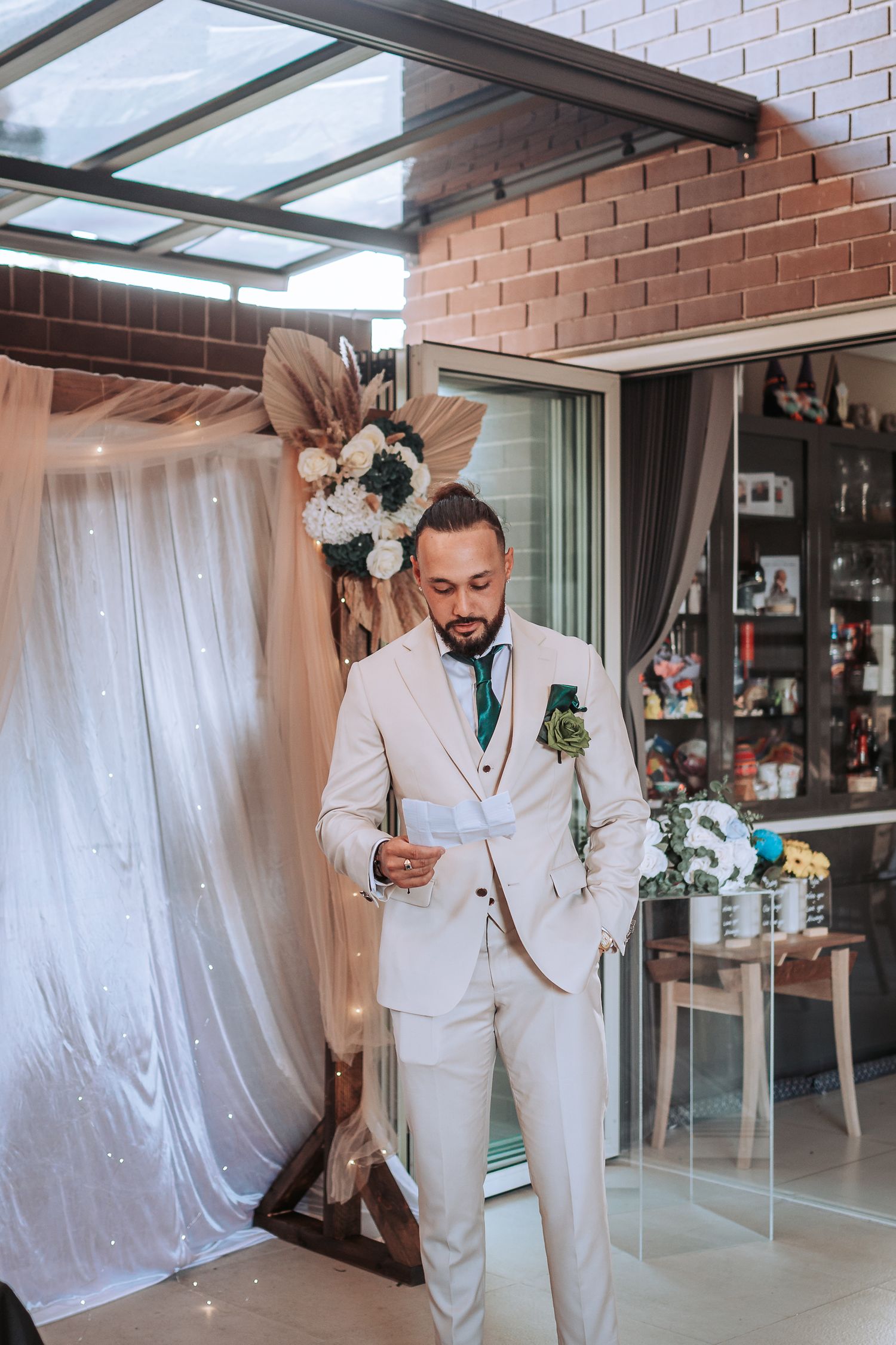 Groom in white suit holding drink against white drapes and floral backdrop.