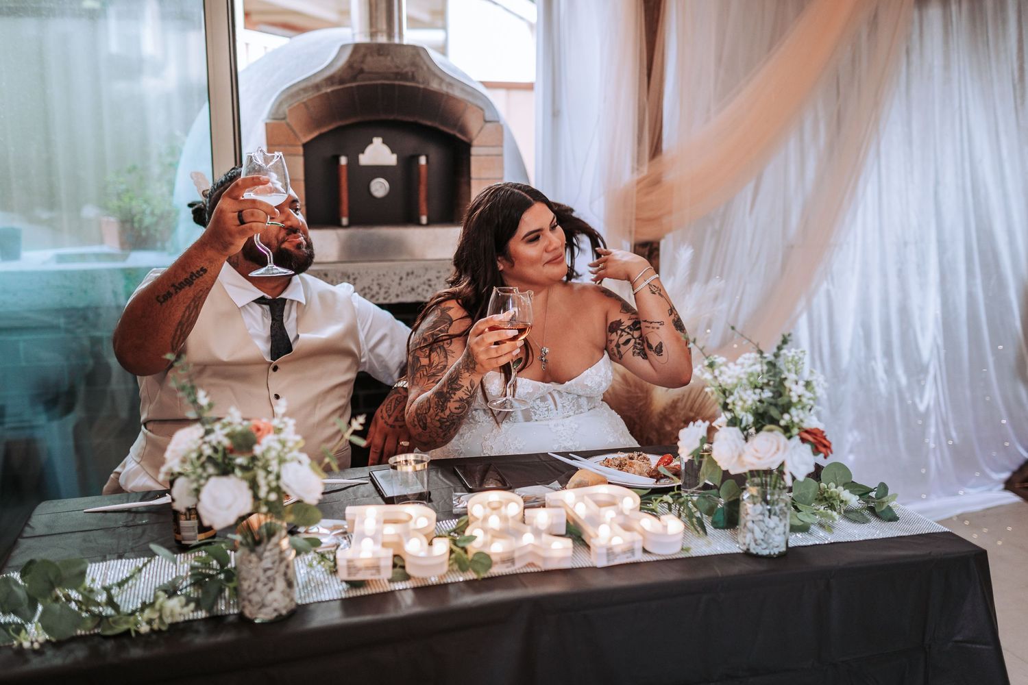 A couple raises their glasses in celebration at their wedding reception table decorated with white flowers and candles.