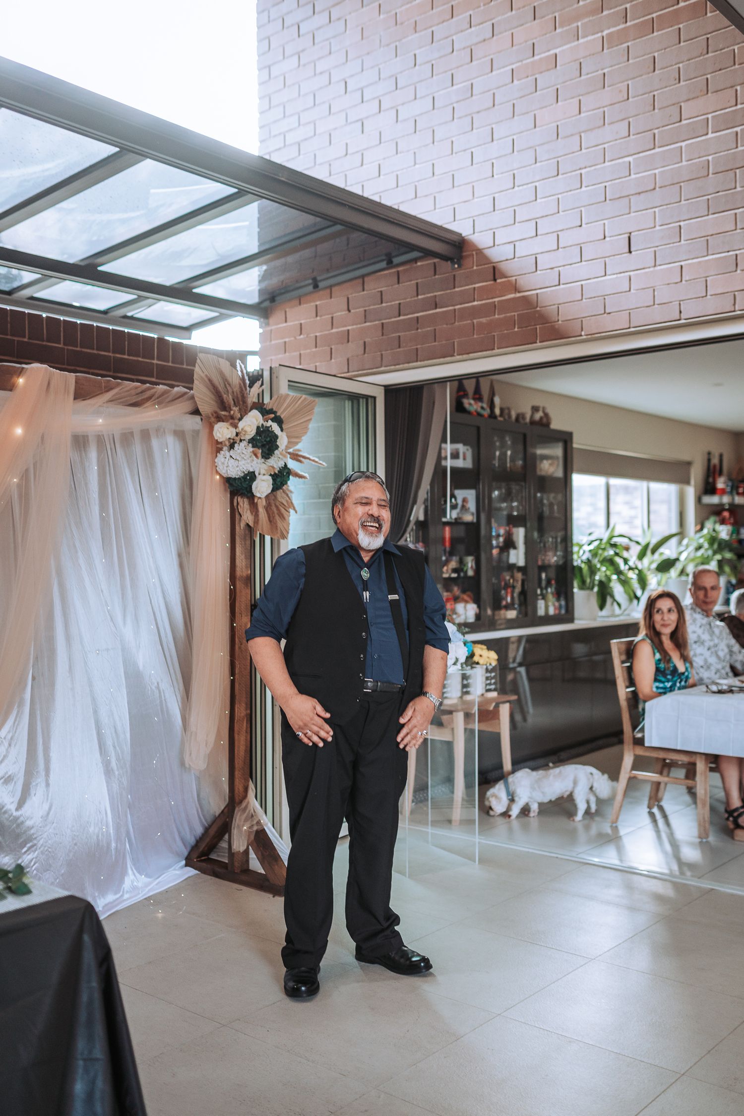 A wedding guest stands in formal attire against brick walls and white draping at an indoor reception.