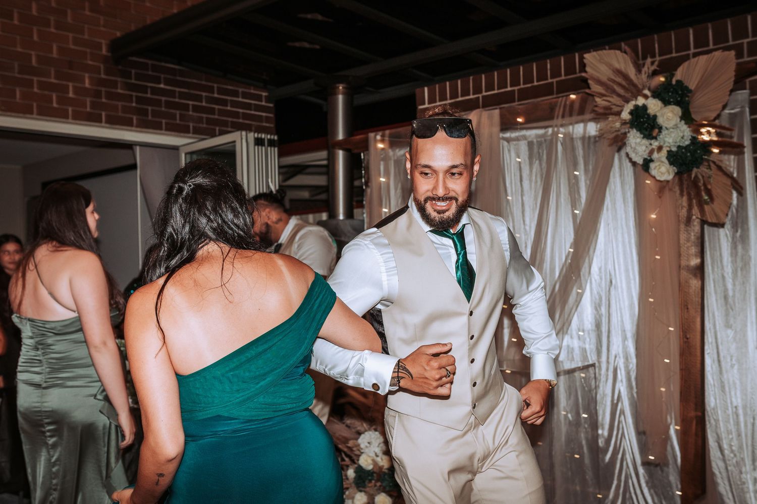 Wedding guests dancing and celebrating at an indoor reception with brick walls and white decor.