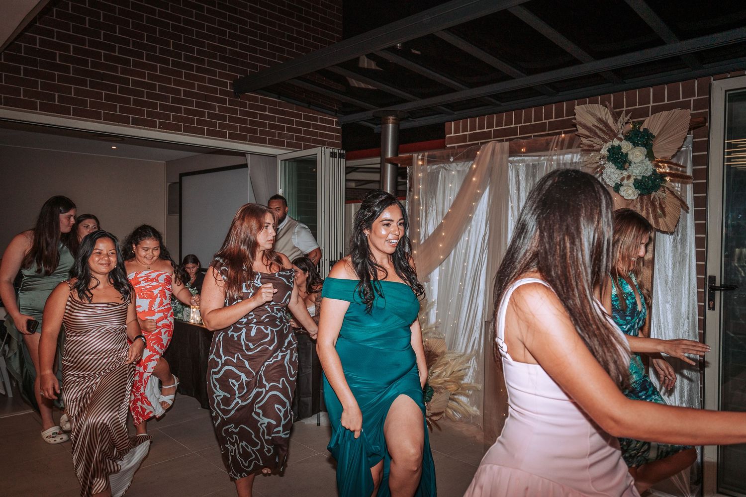 Group of wedding guests dancing in elegant dresses at an indoor reception venue.