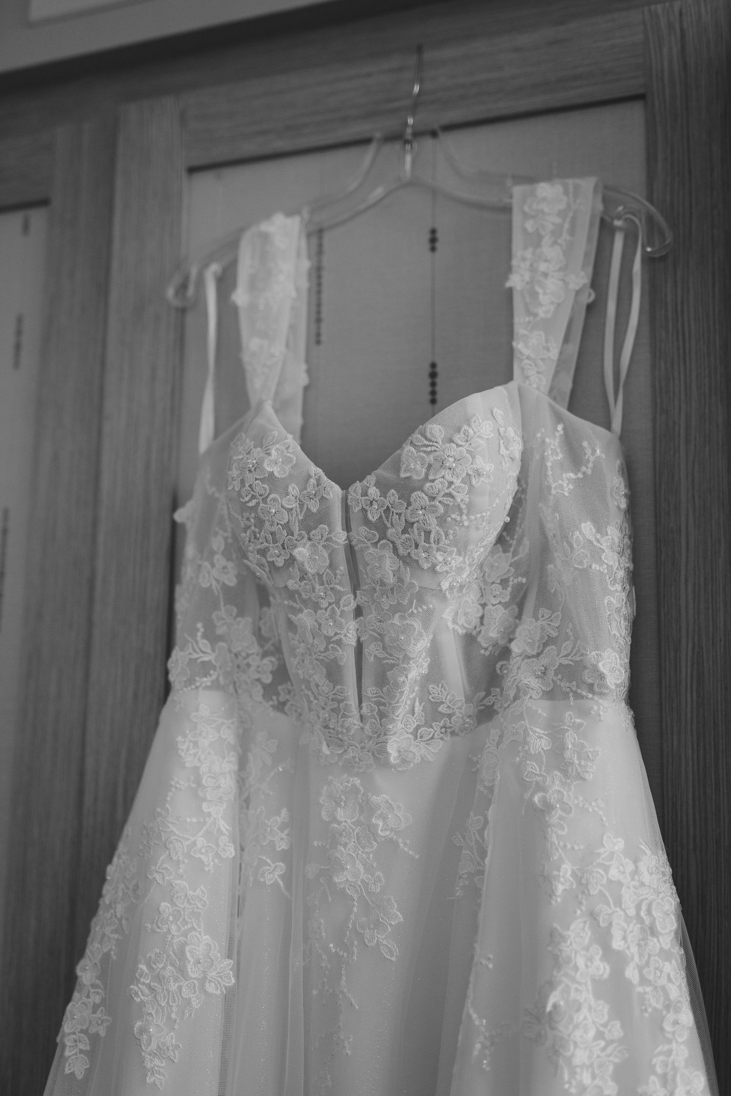 A delicate lace wedding dress with intricate floral details hangs against a wooden door.