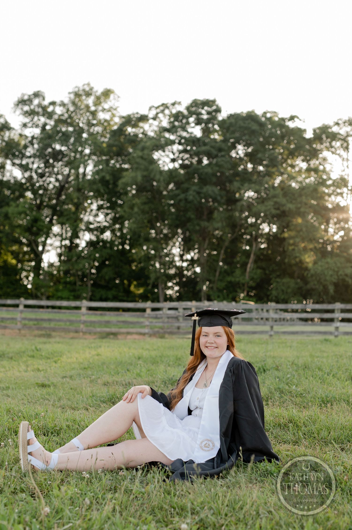 Graduate relaxes on grass wearing white dress and black graduation gown in fenced pasture.