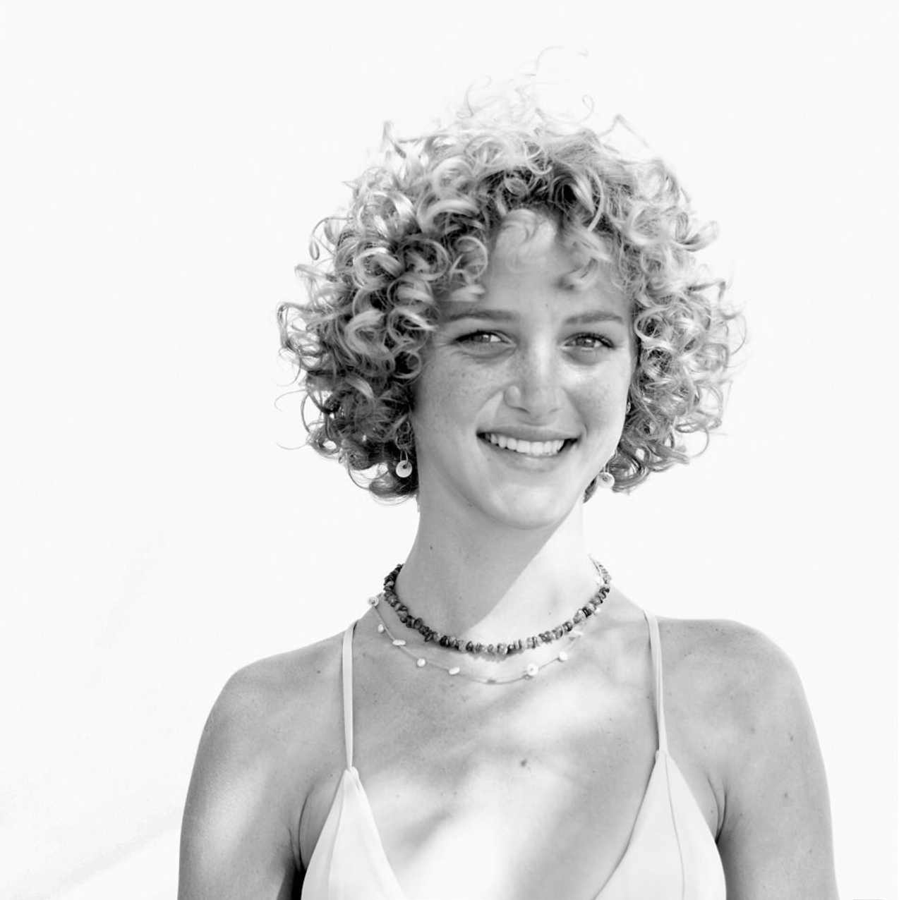 Black and white portrait of a person with curly hair and a beaming smile wearing a necklace against a light background.