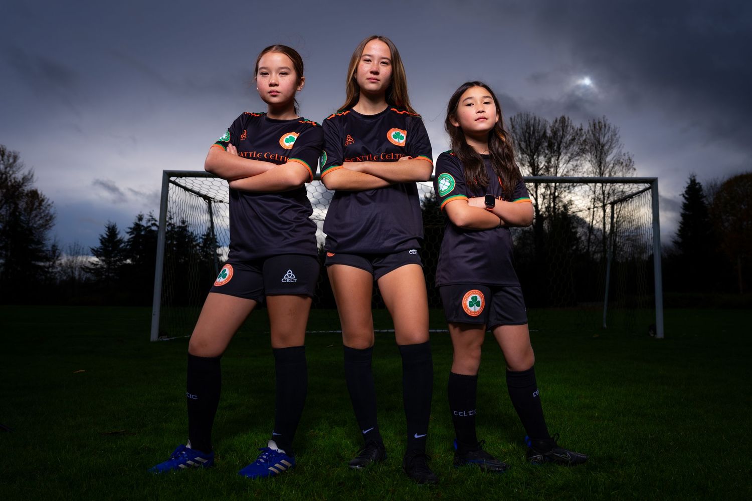 Three soccer players pose confidently in dark uniforms on a field at dusk with a goal visible behind them.