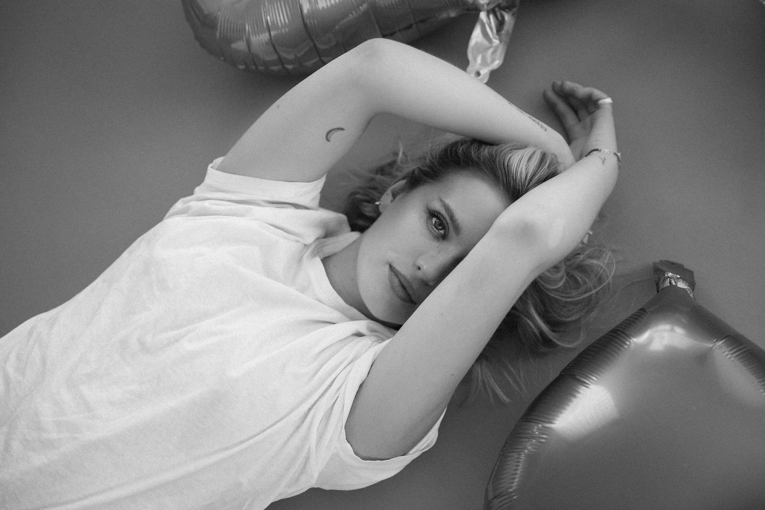 Black and white photo of young woman lying on floor with arms raised above head near balloons.
