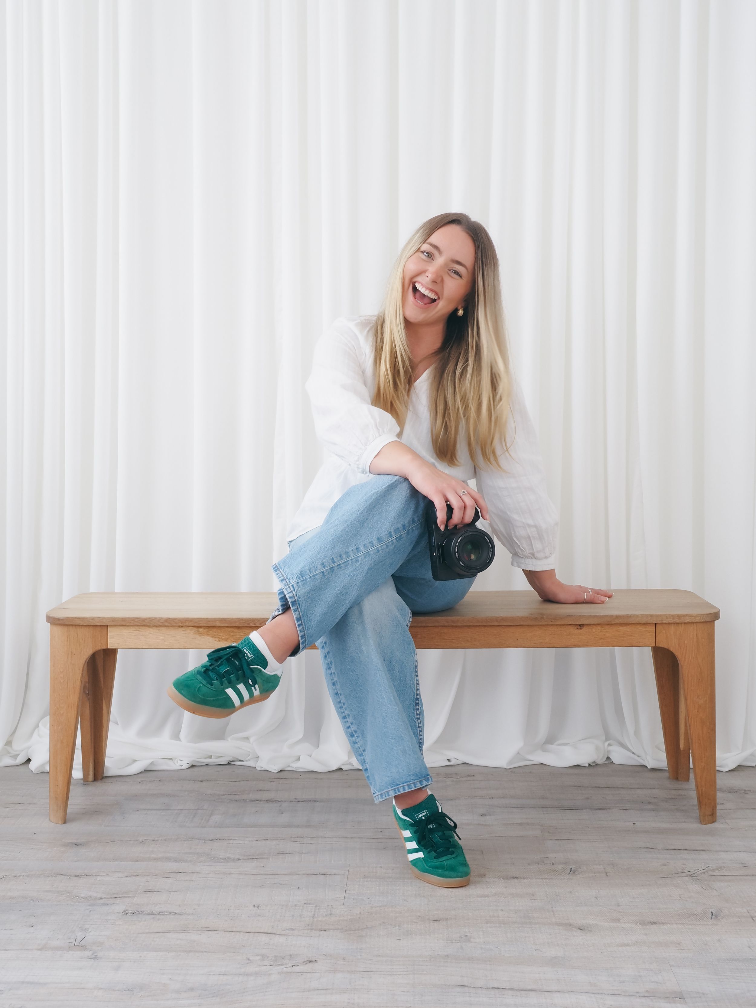 Woman in casual denim outfit sitting on wooden bench against white curtain backdrop.