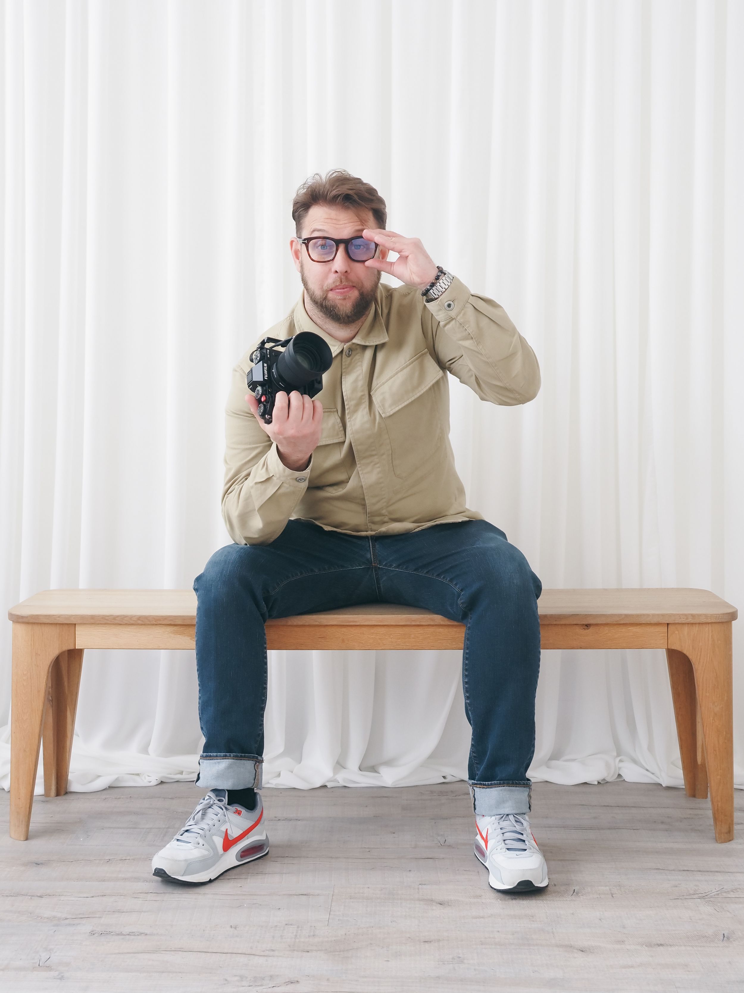 Photographer sitting on wooden bench holding camera in casual attire against white background.