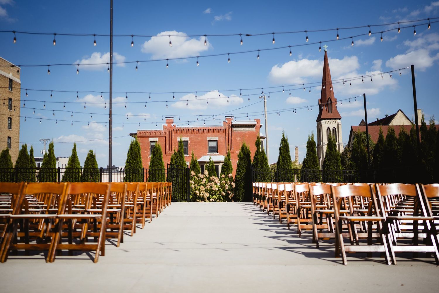 Rows of wooden chairs arranged for an outdoor rooftop wedding ceremony with string lights and city buildings in background.