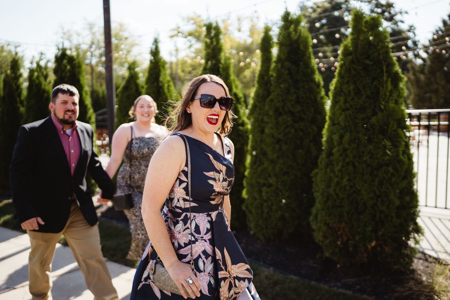 People walking outdoors on a sunny day along a path lined with tall evergreen trees.