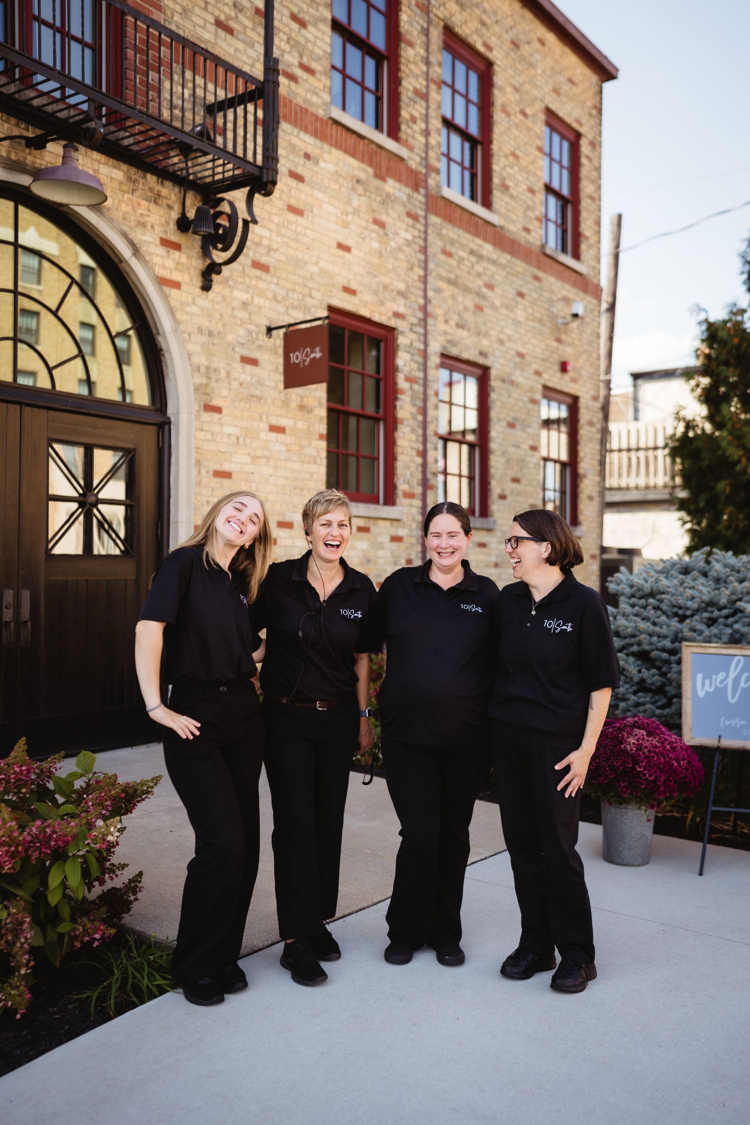 A group of people in black formal attire pose together outside a brick building with arched windows.