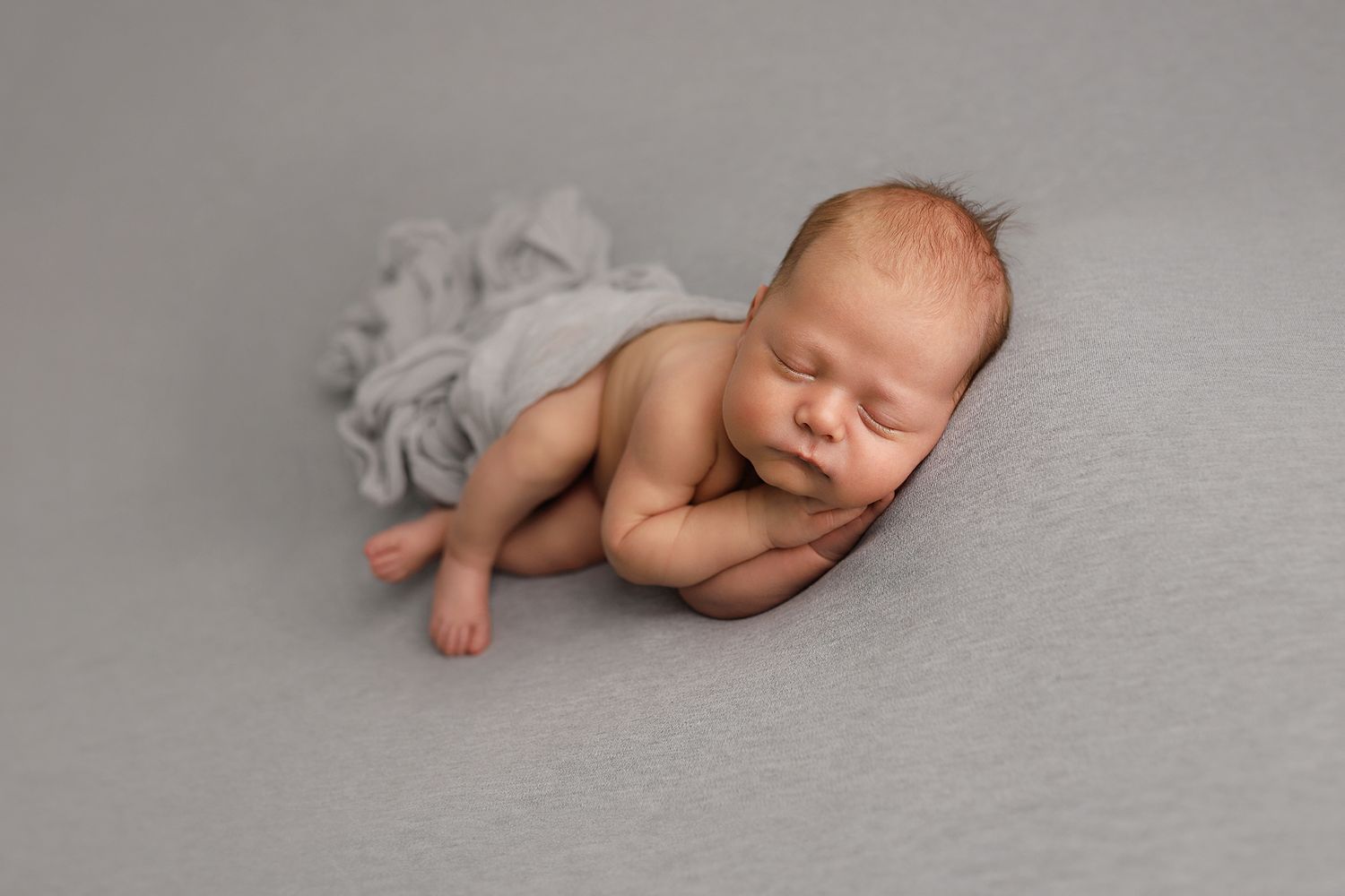 A sleeping newborn baby in a curled up pose on a soft grey backdrop.