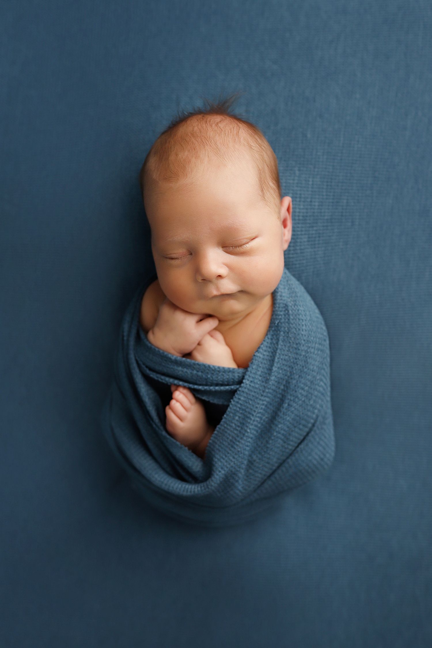 Newborn baby wrapped in soft blue fabric against navy background in artistic sleeping portrait series.