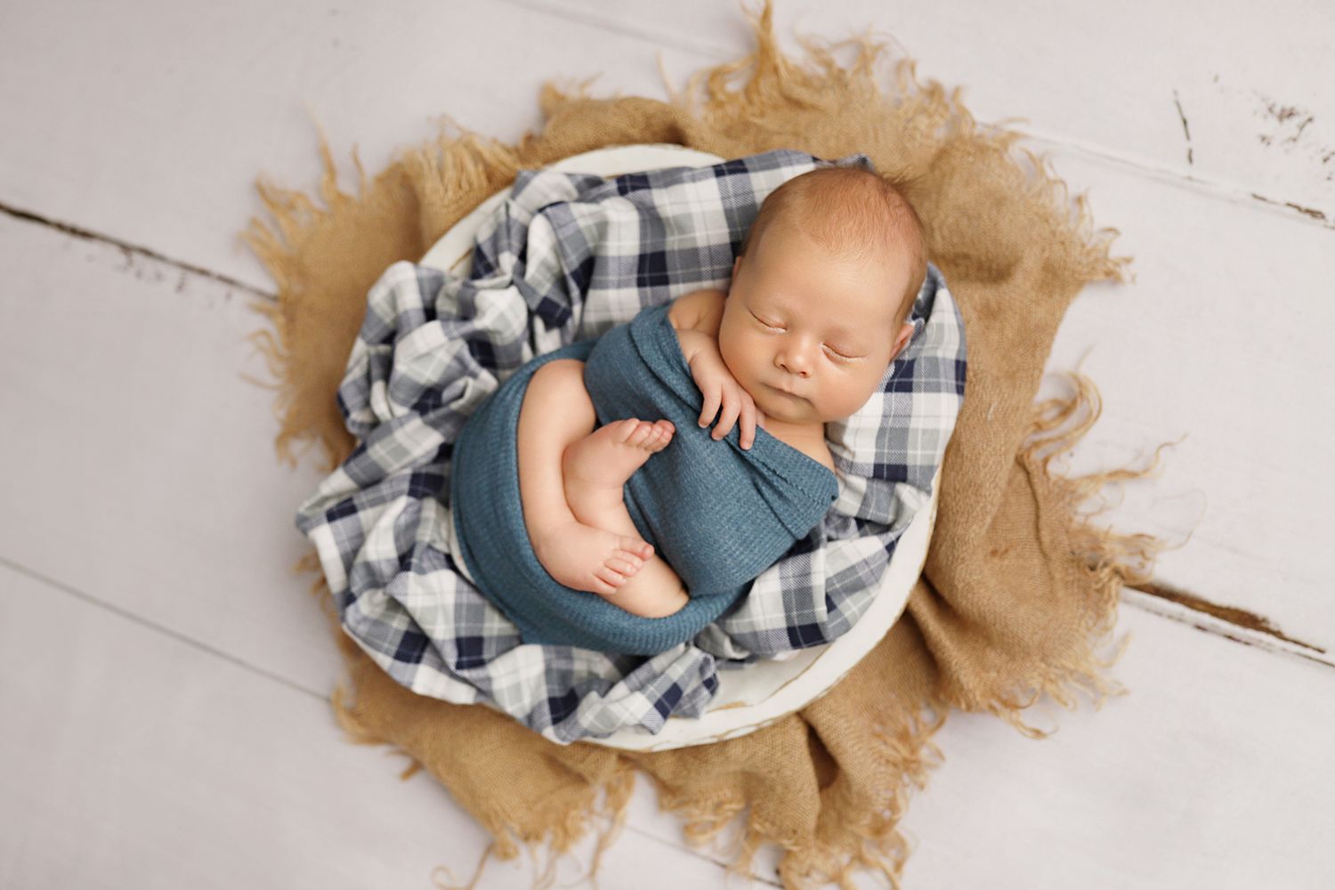 Sleeping newborn baby wearing blue outfit nestled in plaid blanket on rustic wooden floor with burlap accents.