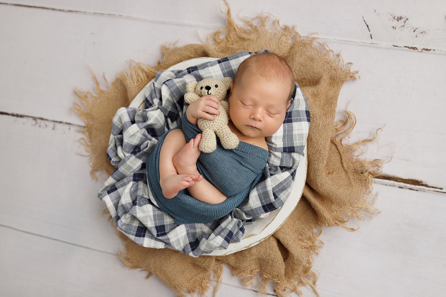 Newborn baby wrapped in plaid blanket lying on rustic burlap fabric against white wooden background.