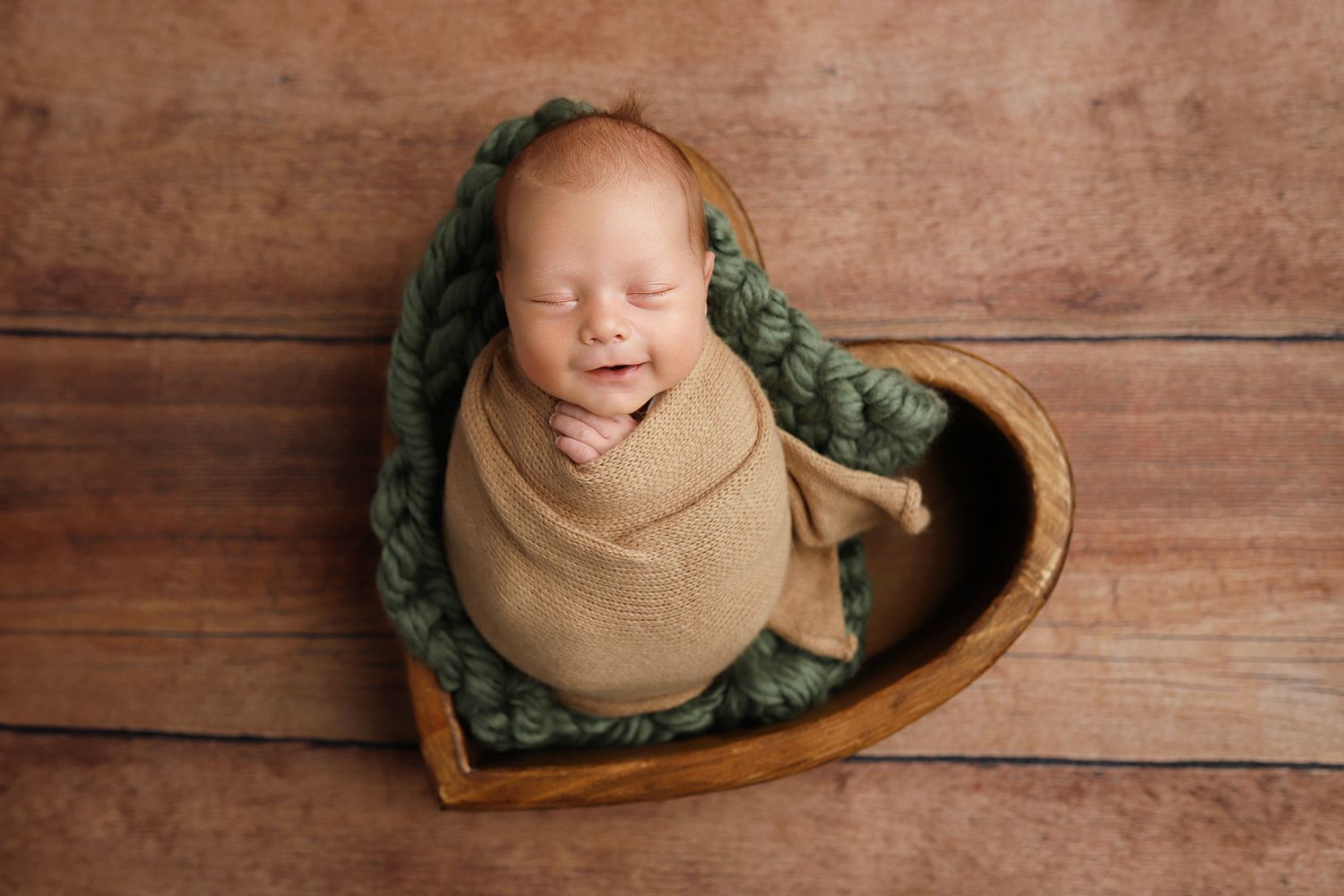 Sleeping newborn wrapped in beige swaddle rests in wooden heart bowl with green blanket on rustic wood floor.