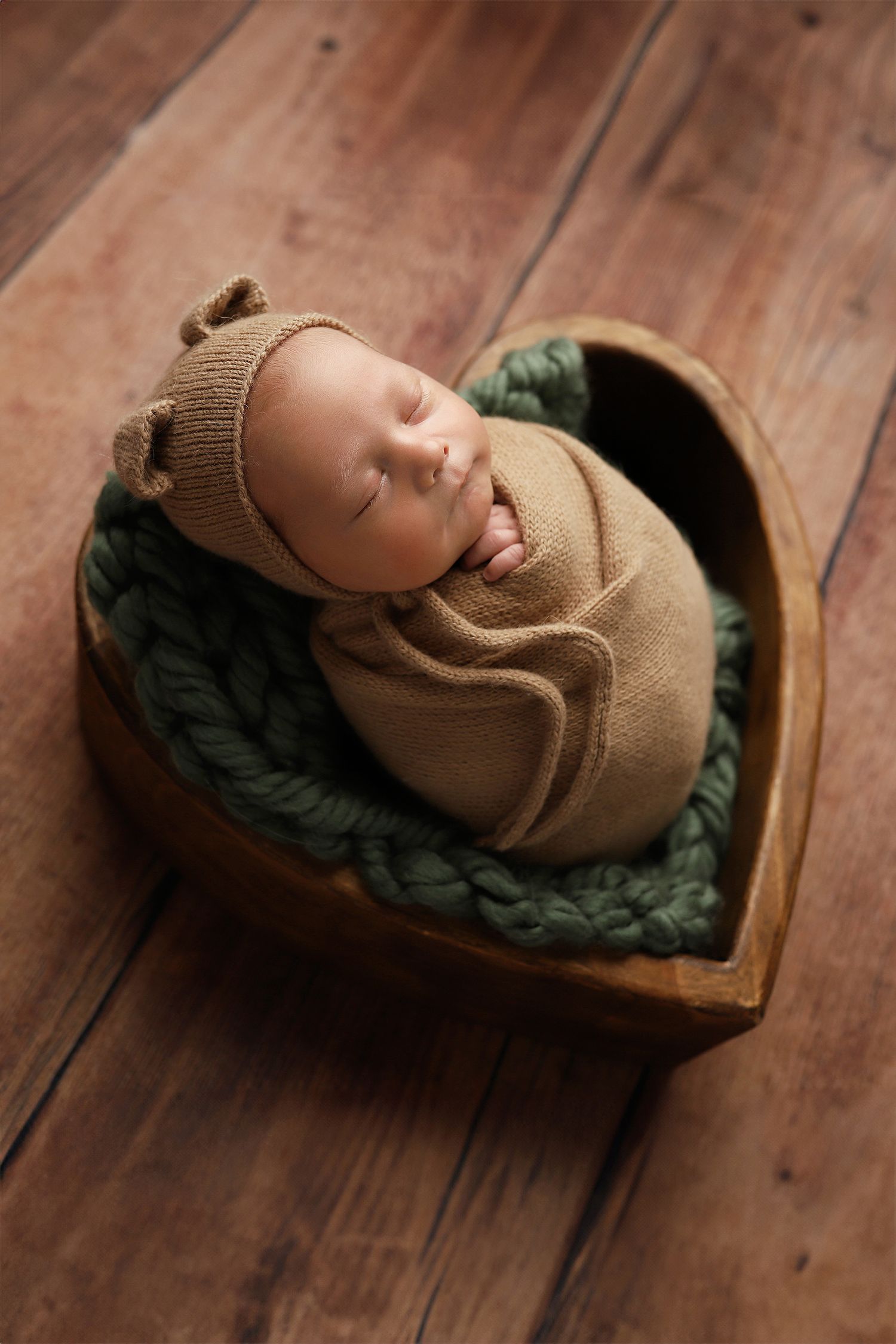 Newborn wrapped in beige knit swaddle and hat sleeping peacefully in wooden heart-shaped basket with green padding.