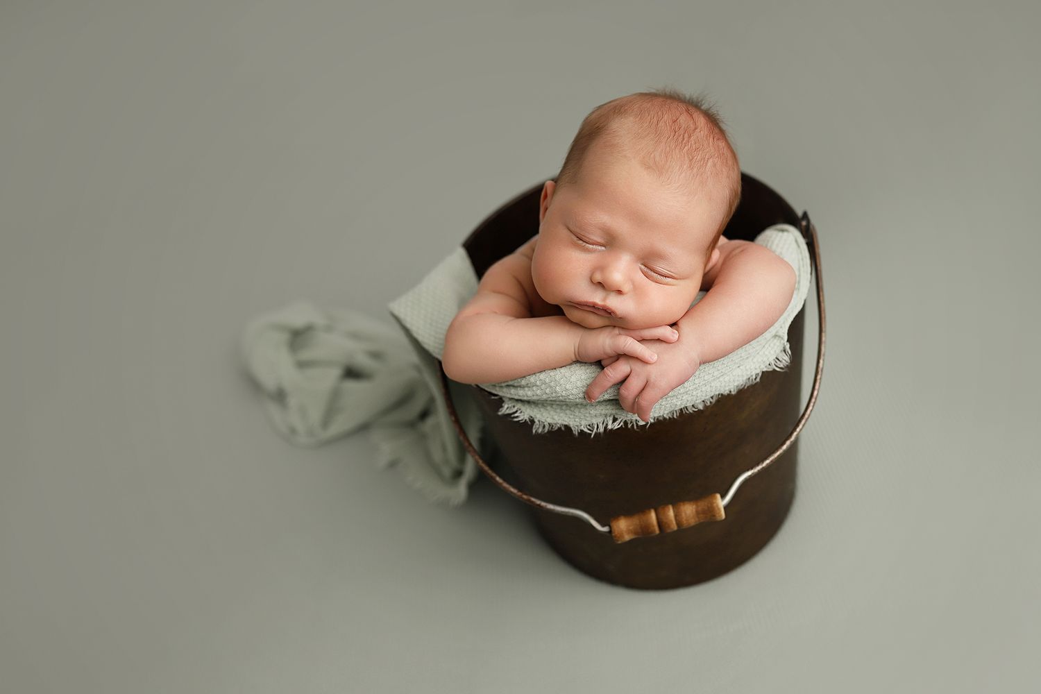 A sleeping newborn baby nestled in a cozy brown wooden bowl against a neutral gray backdrop.