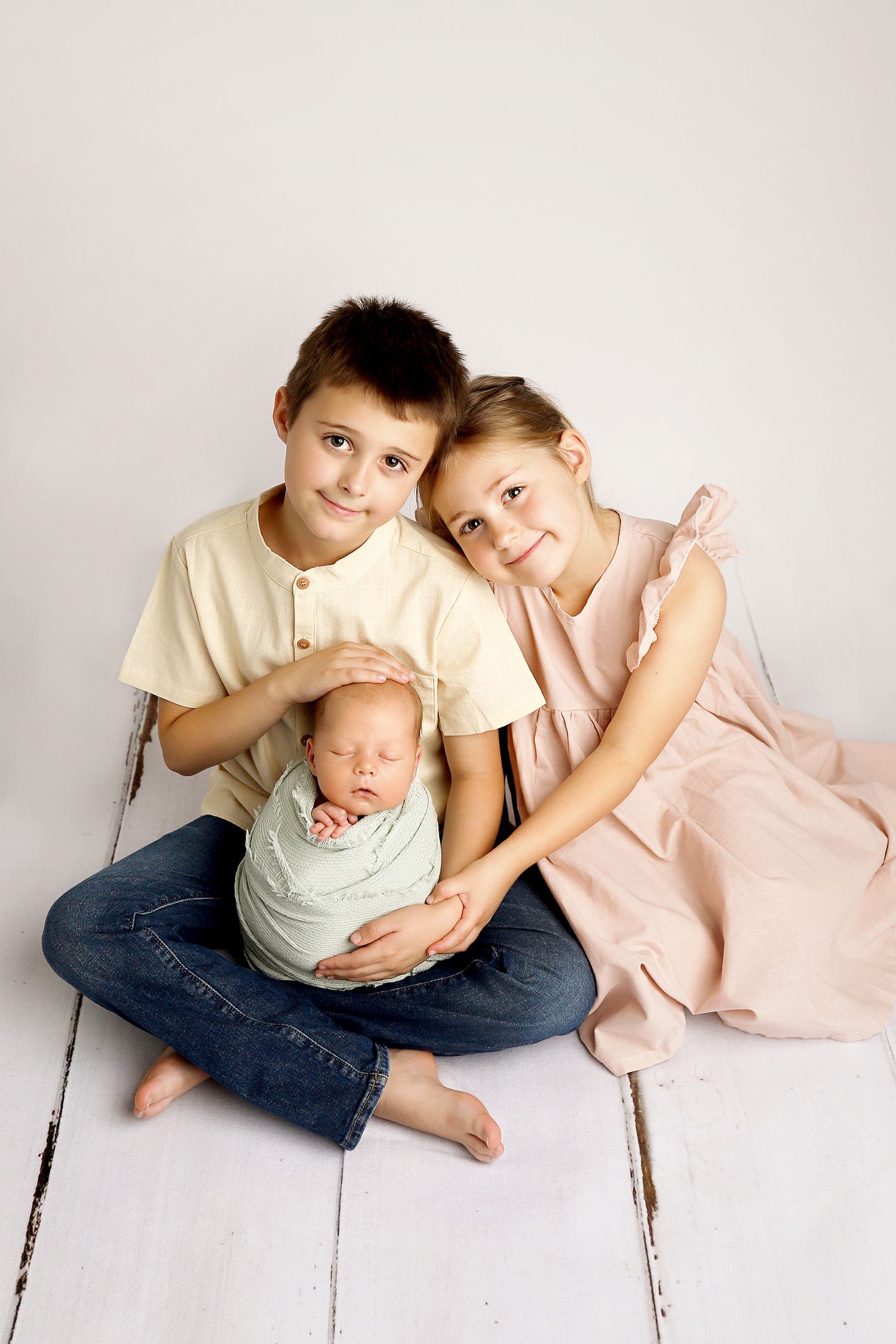 A heartwarming family portrait series showing siblings bonding with a newborn baby on a white wooden floor.