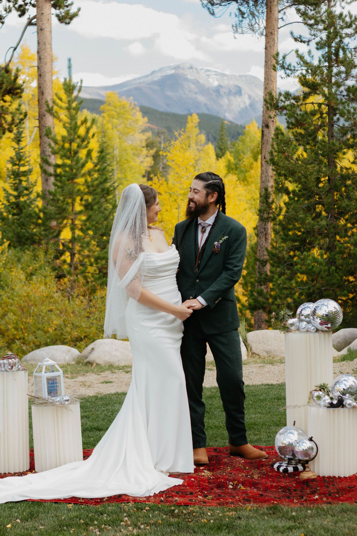 A couple shares a tender moment during their outdoor mountain wedding ceremony against a backdrop of fall foliage and evergreens.