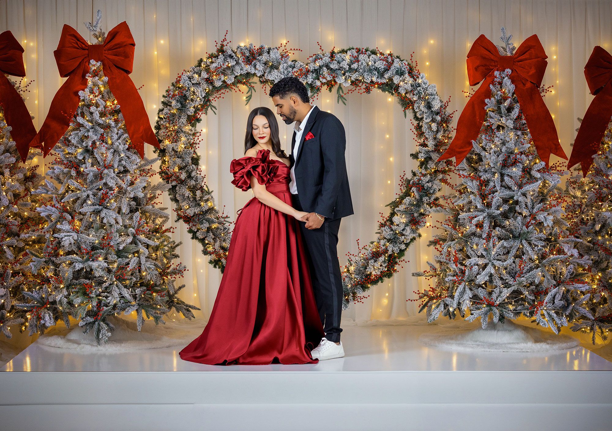 Couple in formal attire poses in front of decorated Christmas trees with silver and white ornaments and twinkling lights.