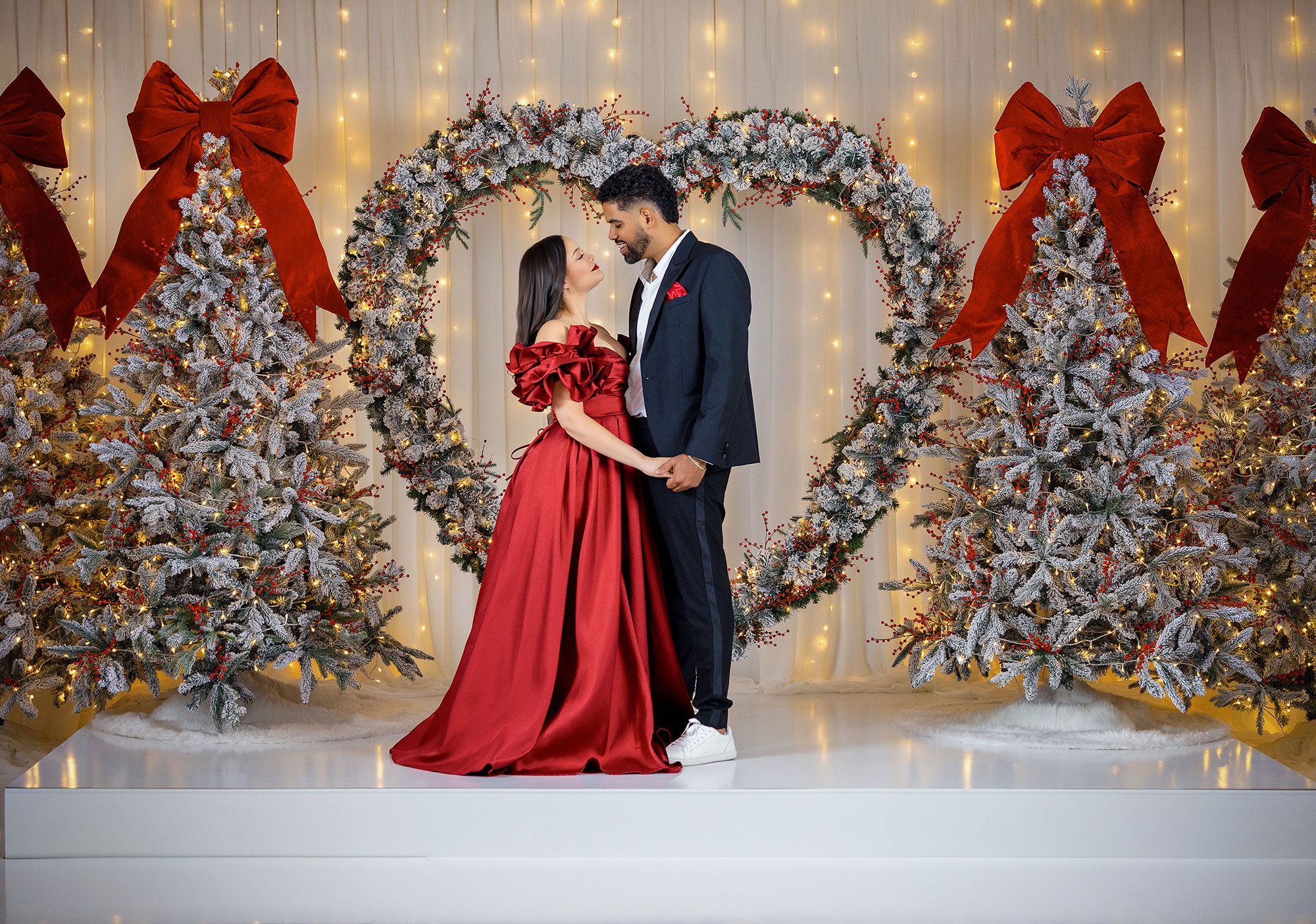 Couple poses in front of a festive holiday backdrop with white lights and decorated Christmas trees.